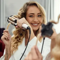 Uśmiechnięta blondynka tworzy piękne fale na włosach za pomocą lokówki.