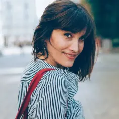 Uśmiechnięta kobieta po czterdziestce z modnymi fryzurami z grzywką, w pasiastym ubraniu i z czerwoną torbą.